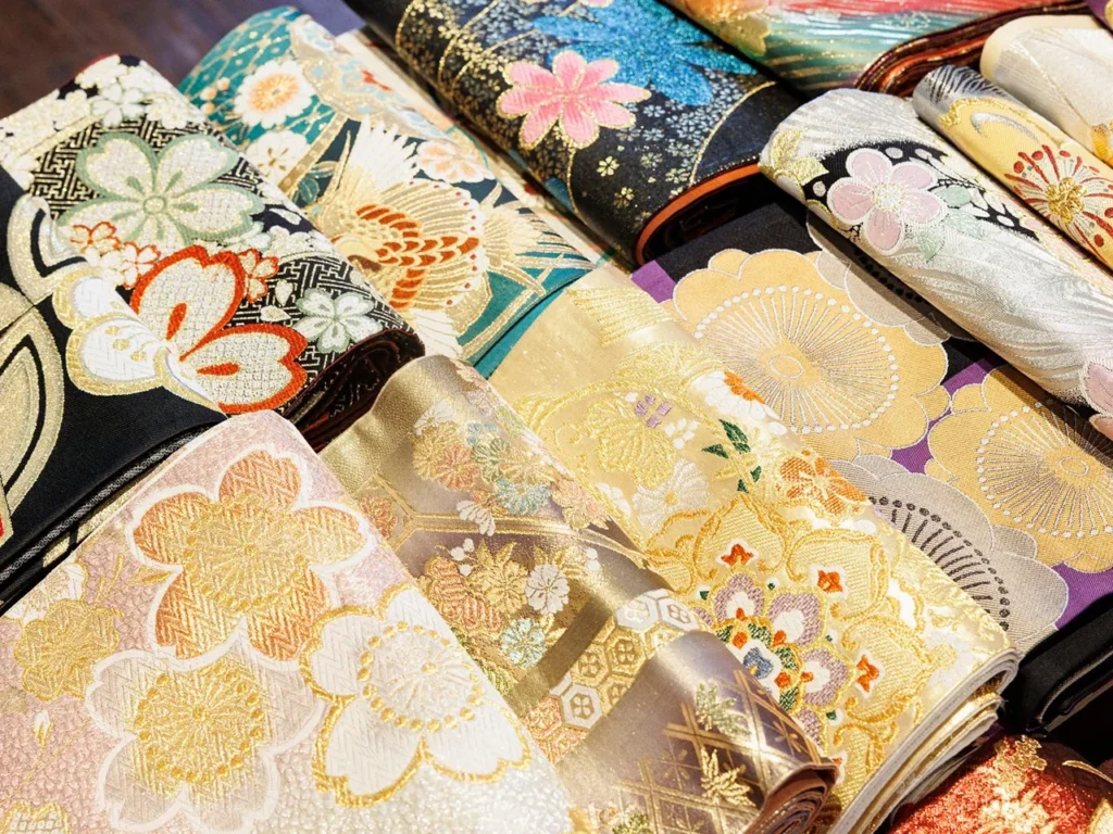桜や鶴、古典柄が金糸で豪華に織り込まれた、色とりどりの美しい和装生地の反物が並ぶ様子。