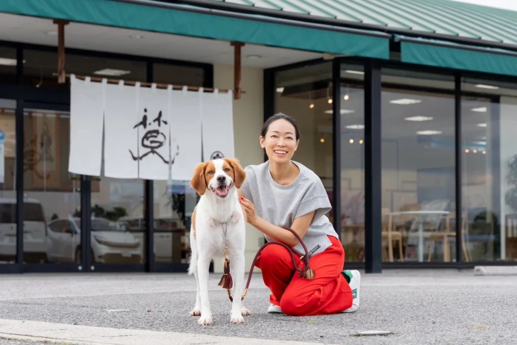 小倉の暖簾があるANNACANEの店舗前で、代表の吉永 澪叶がリードを付けた犬に膝をついて寄り添っています。